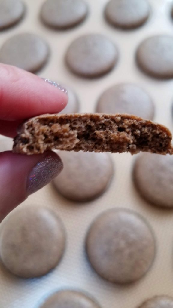 A chocolate macaron shell held up to show its texture, with a tray of baked shells behind