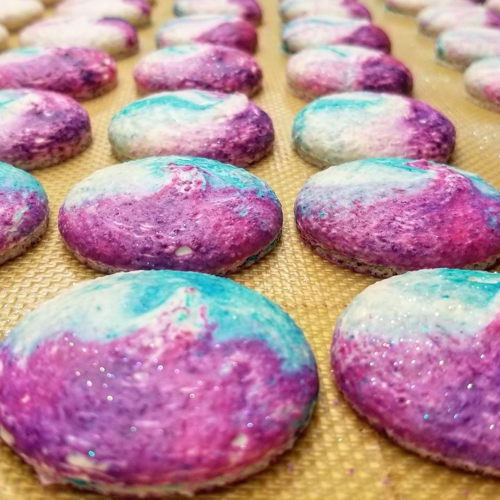 Pink and teal galaxy-swirl macaron shells with glitter on a baking mat
