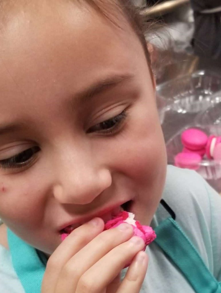 A young baker in an apron happily tasting a pink macaron she made in class